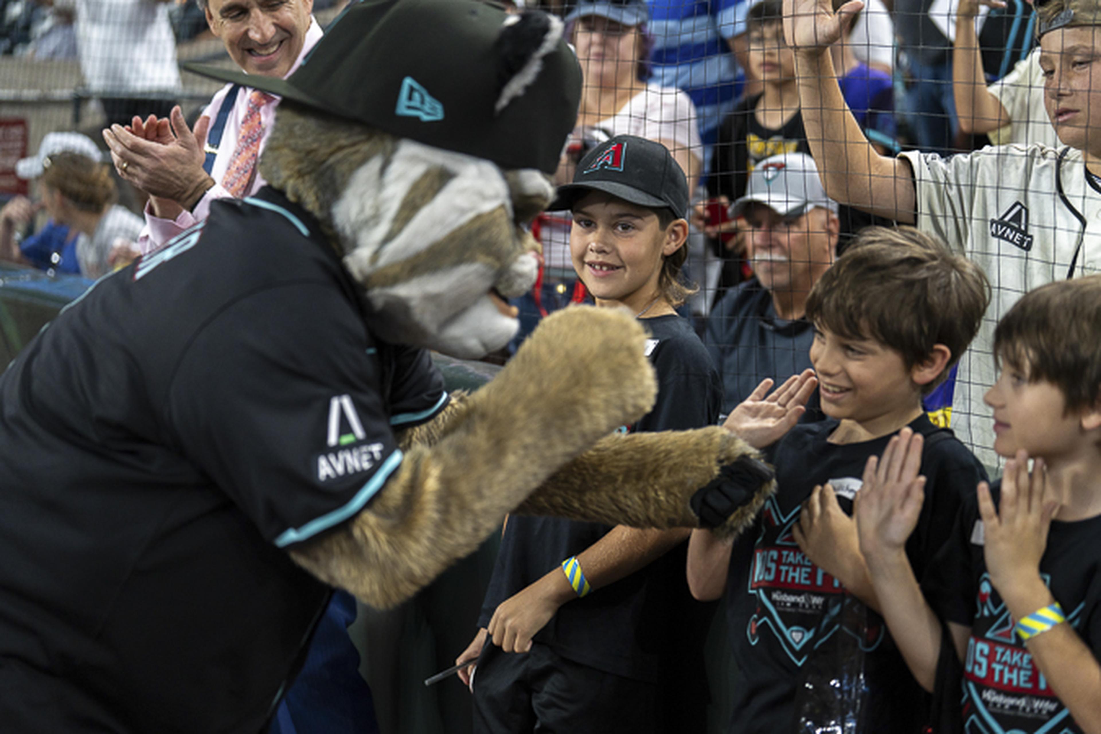 Baxter the Bobcat, wearing a black cap, high‑fives excited kids in matching Diamondbacks T‑shirts at a packed stadium.