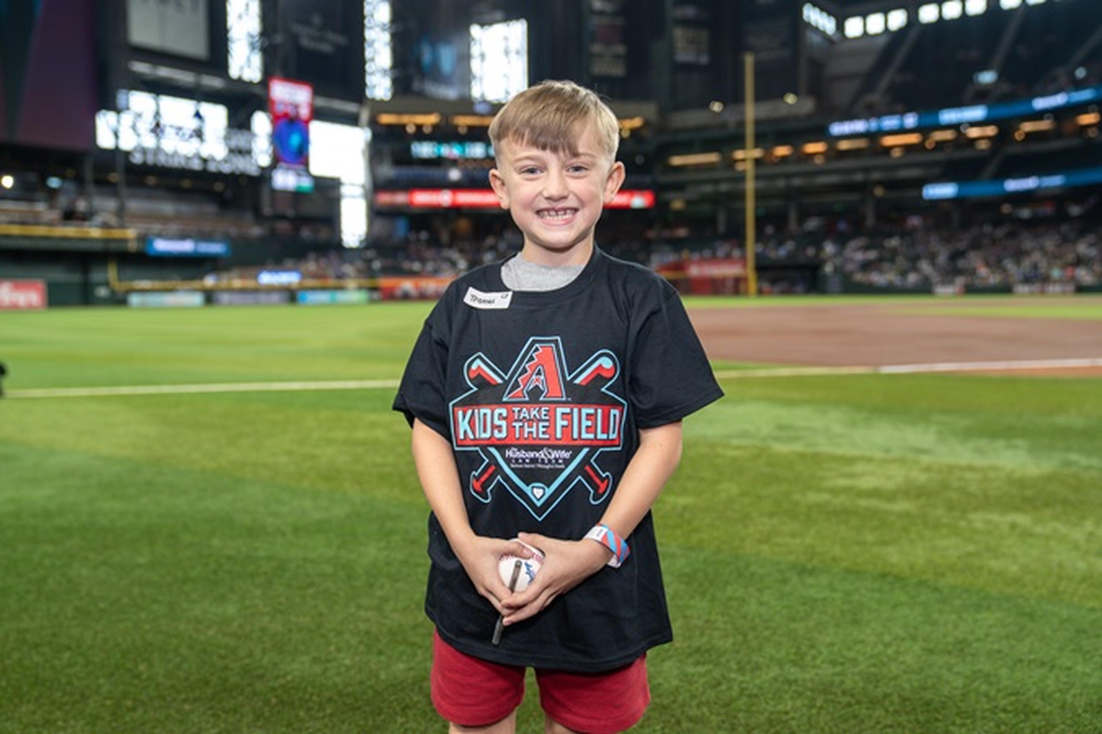 Young boy smiling on a baseball field, wearing a "Kids Take the Field" T-shirt, holding a baseball. Stadium lights and audience in the background.