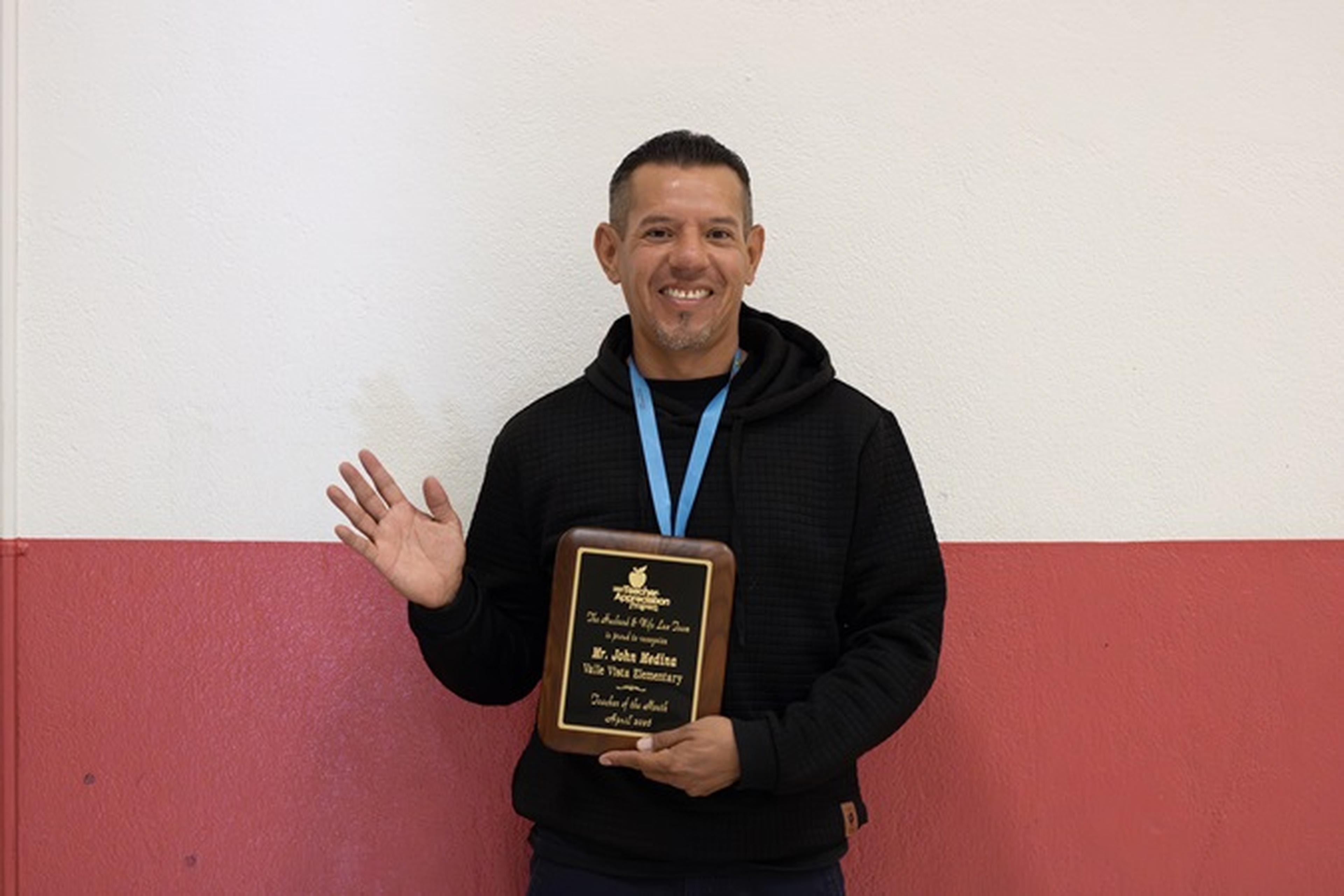 Mr. Medina, this month’s Husband & Wife Law Team Teacher Appreciation Award honoree, stands proudly against a red‑and‑white wall.