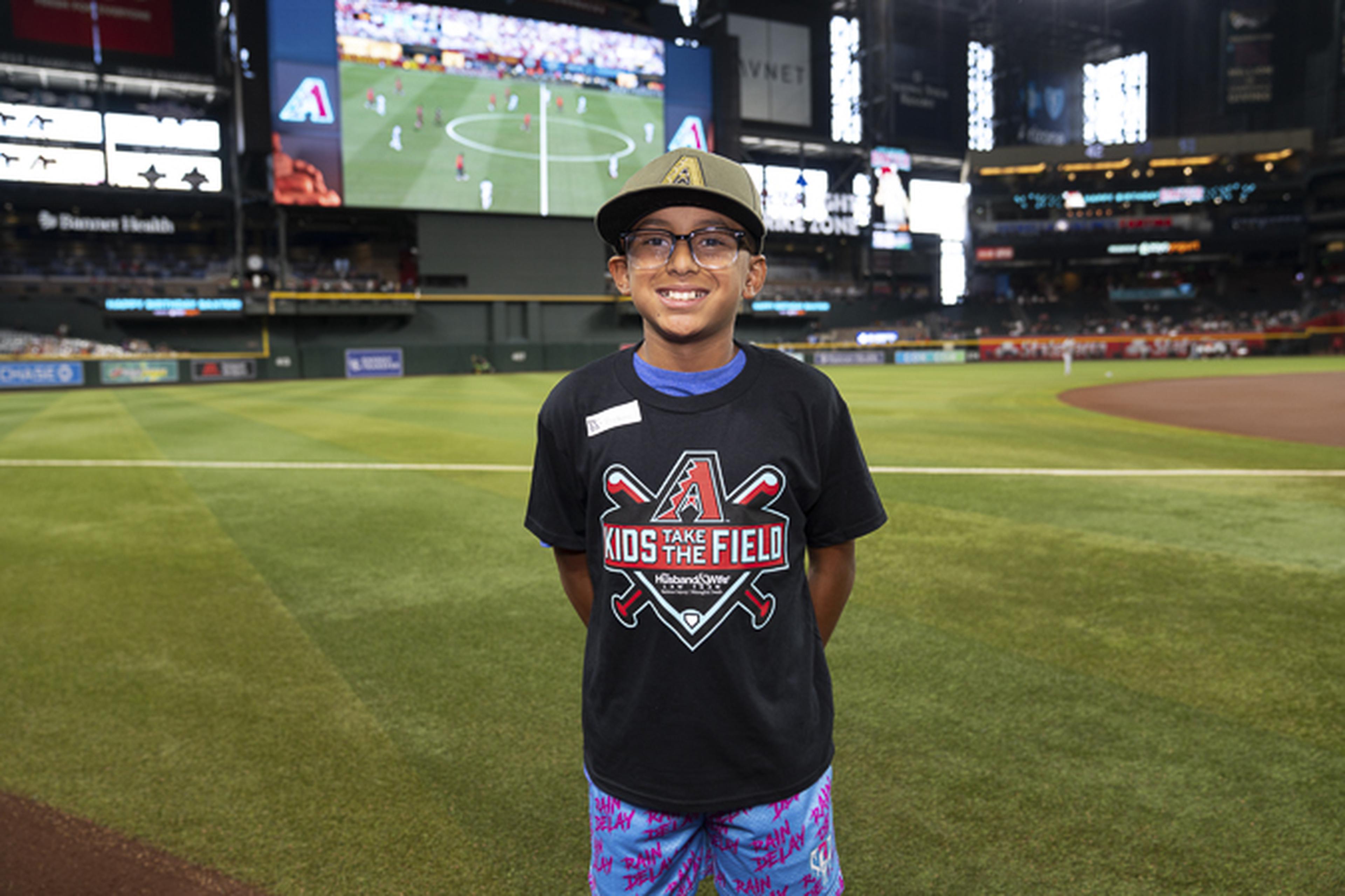 Chasing big-league dreams, this young baseball fan proudly smiles on the field in a Kids Take the Field shirt, making memories that will last a lifetime.