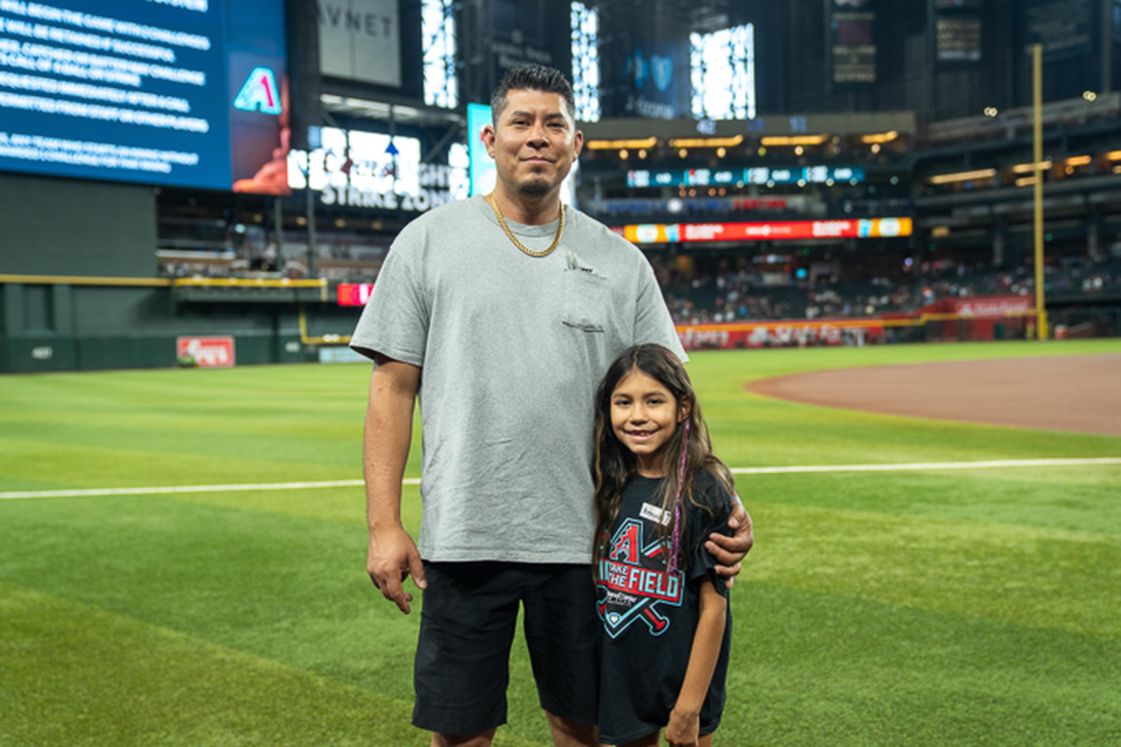 Father and Daughter smile together on a baseball field, with a stadium full of spectators rising behind them.
