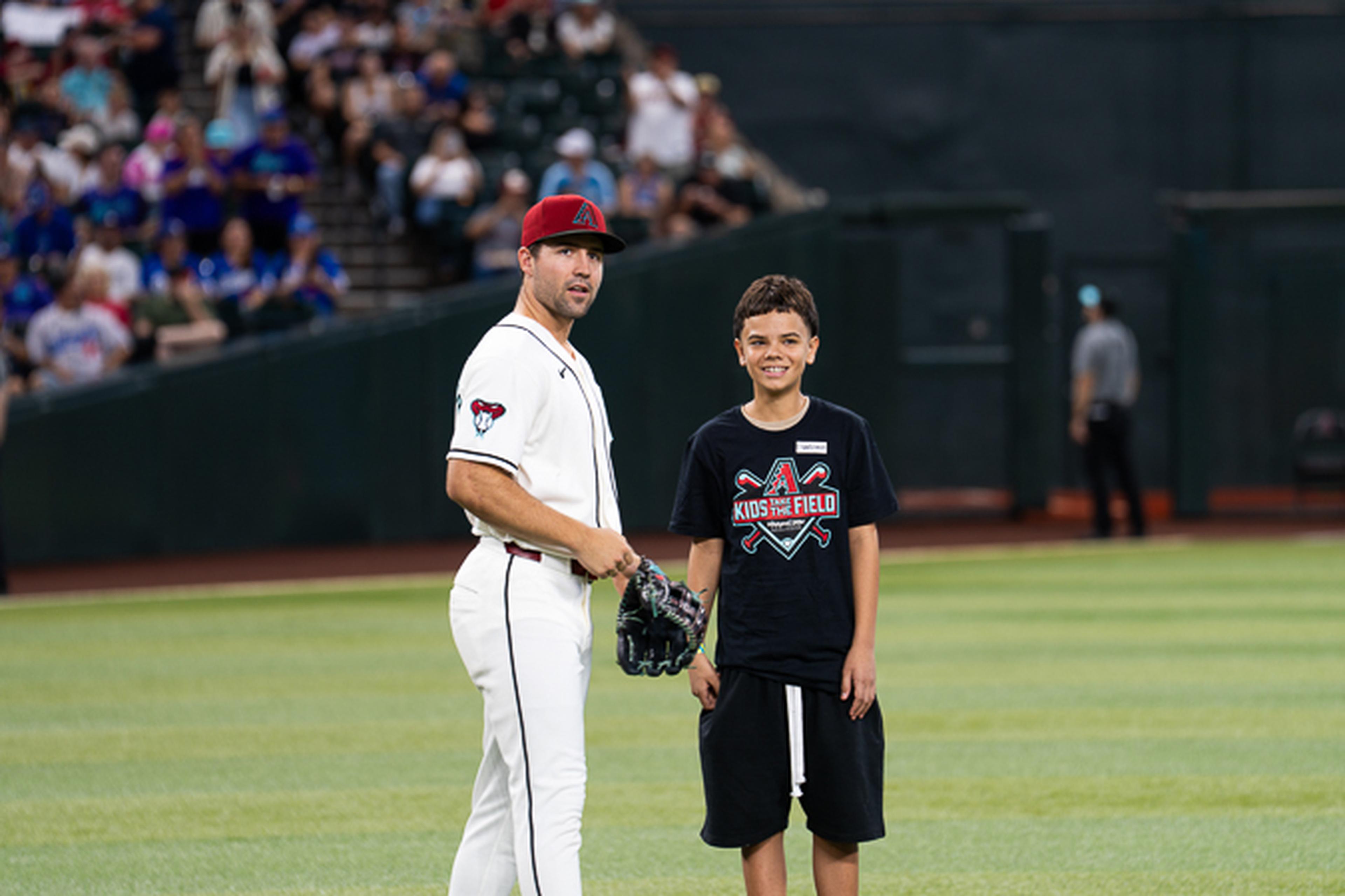 A young Kids Take the Field participant stands alongside an Arizona Diamondbacks player on the field, enjoying a memorable experience in front of a packed crowd at Chase Field.