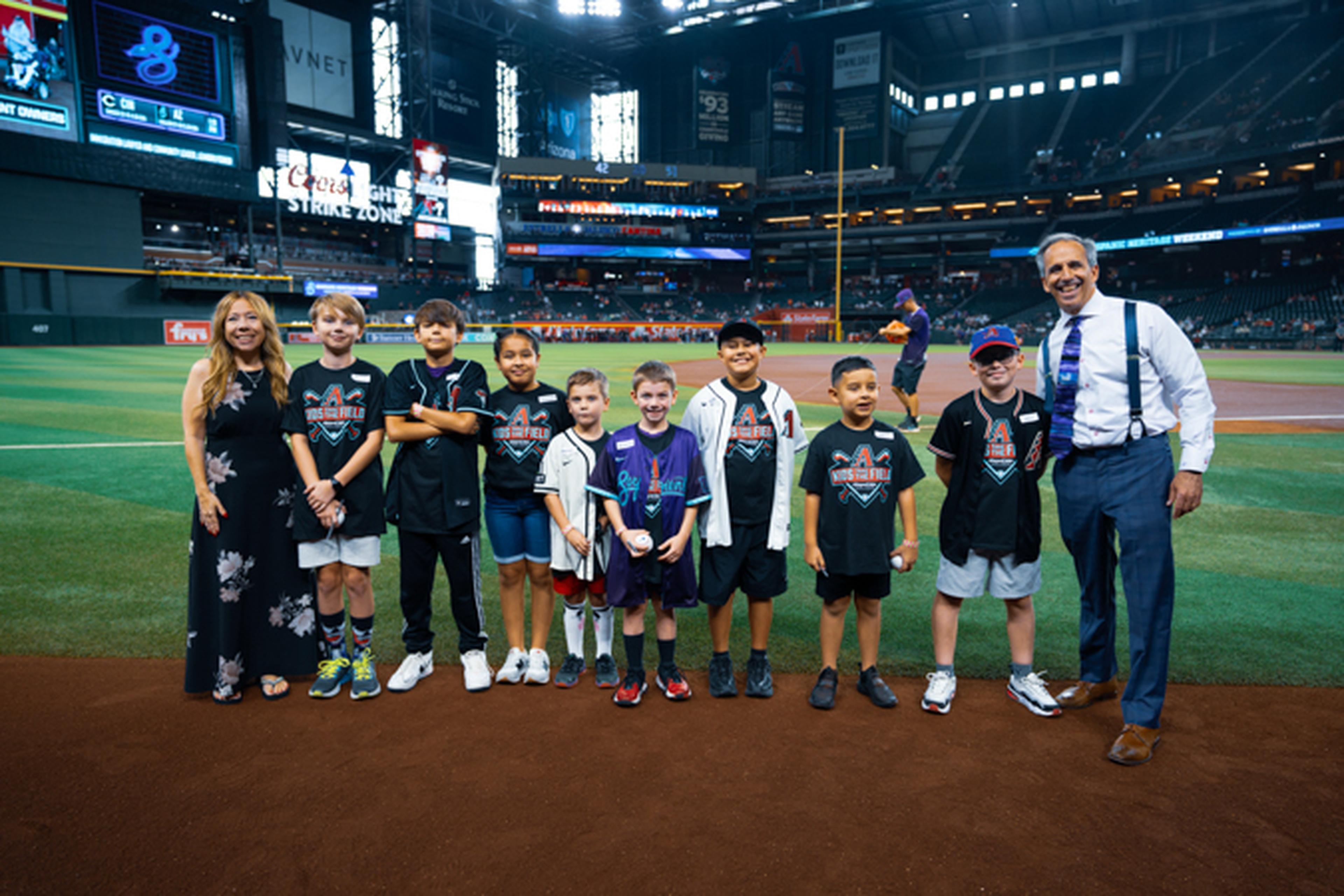 A group of children and two adults stand on a baseball field, smiling. The stadium is well-lit, with visible seating and signage in the background.