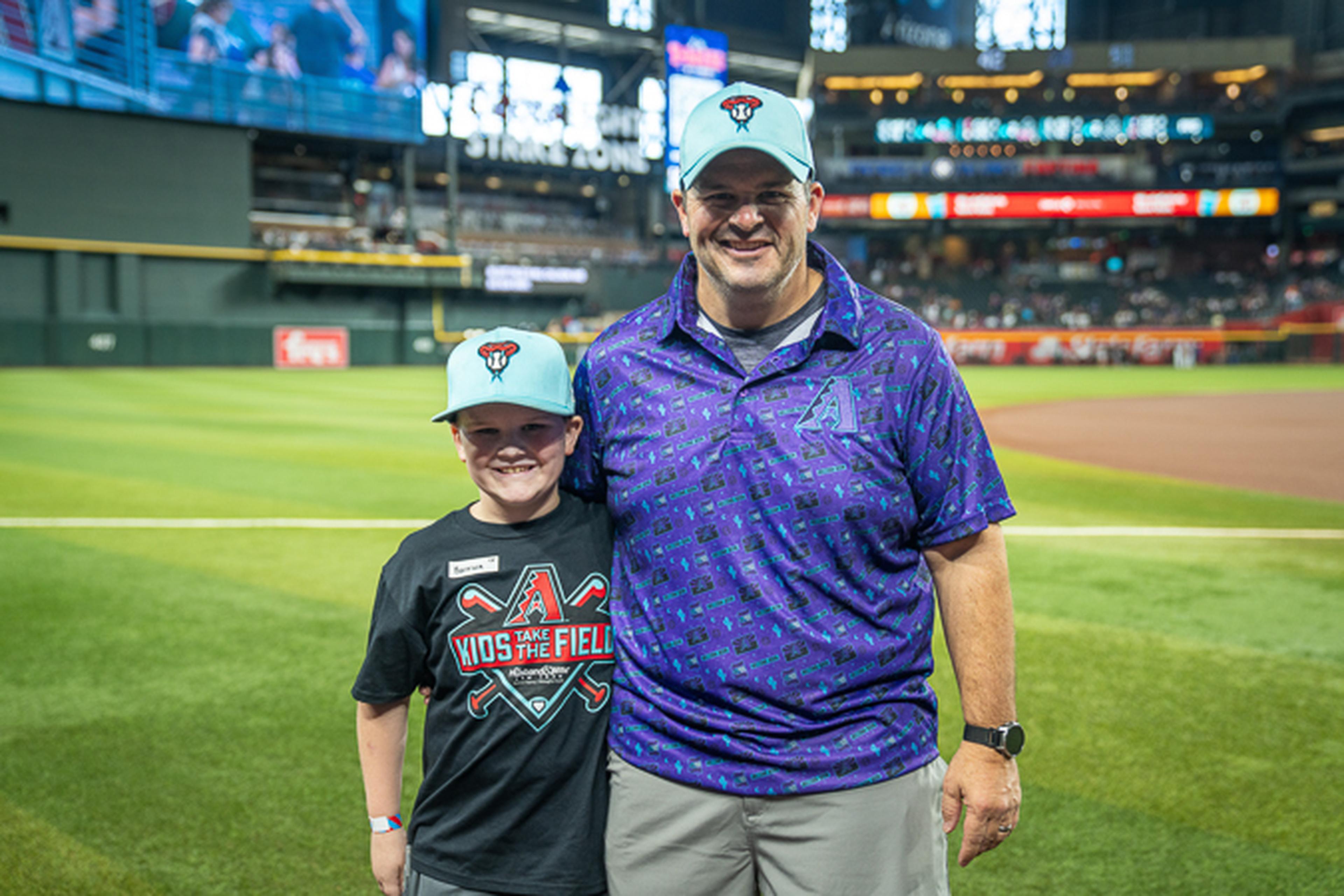 Father and son smile on the baseball field, both wearing matching Arizona Diamondbacks caps and team gear, with the stadium rising behind them.