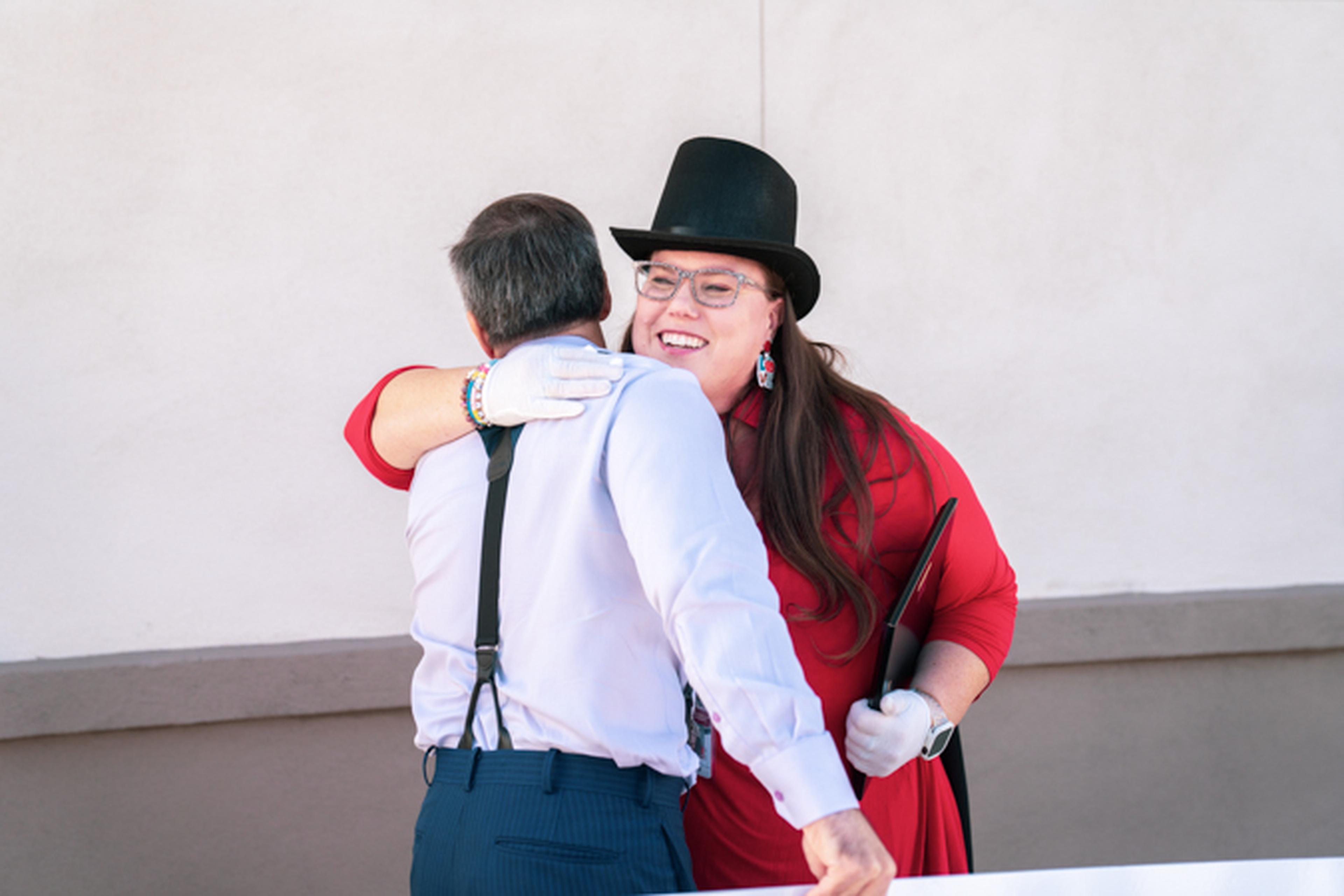 A person in a top hat and red dress hugs another person wearing a white shirt and suspenders against a plain background.