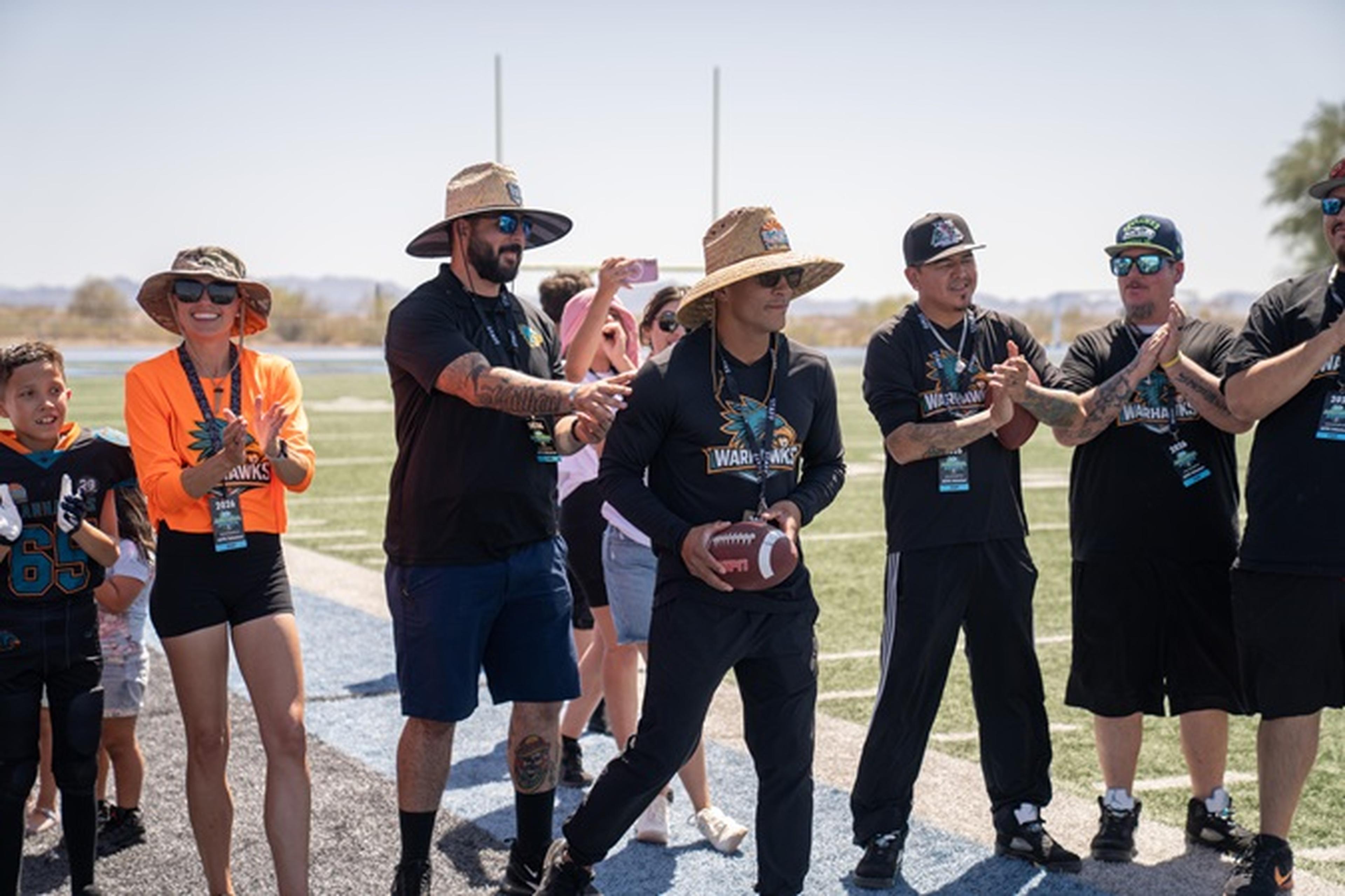 AYFL members in team gear stand on the field as Coach Nino holds a football and the group cheers.