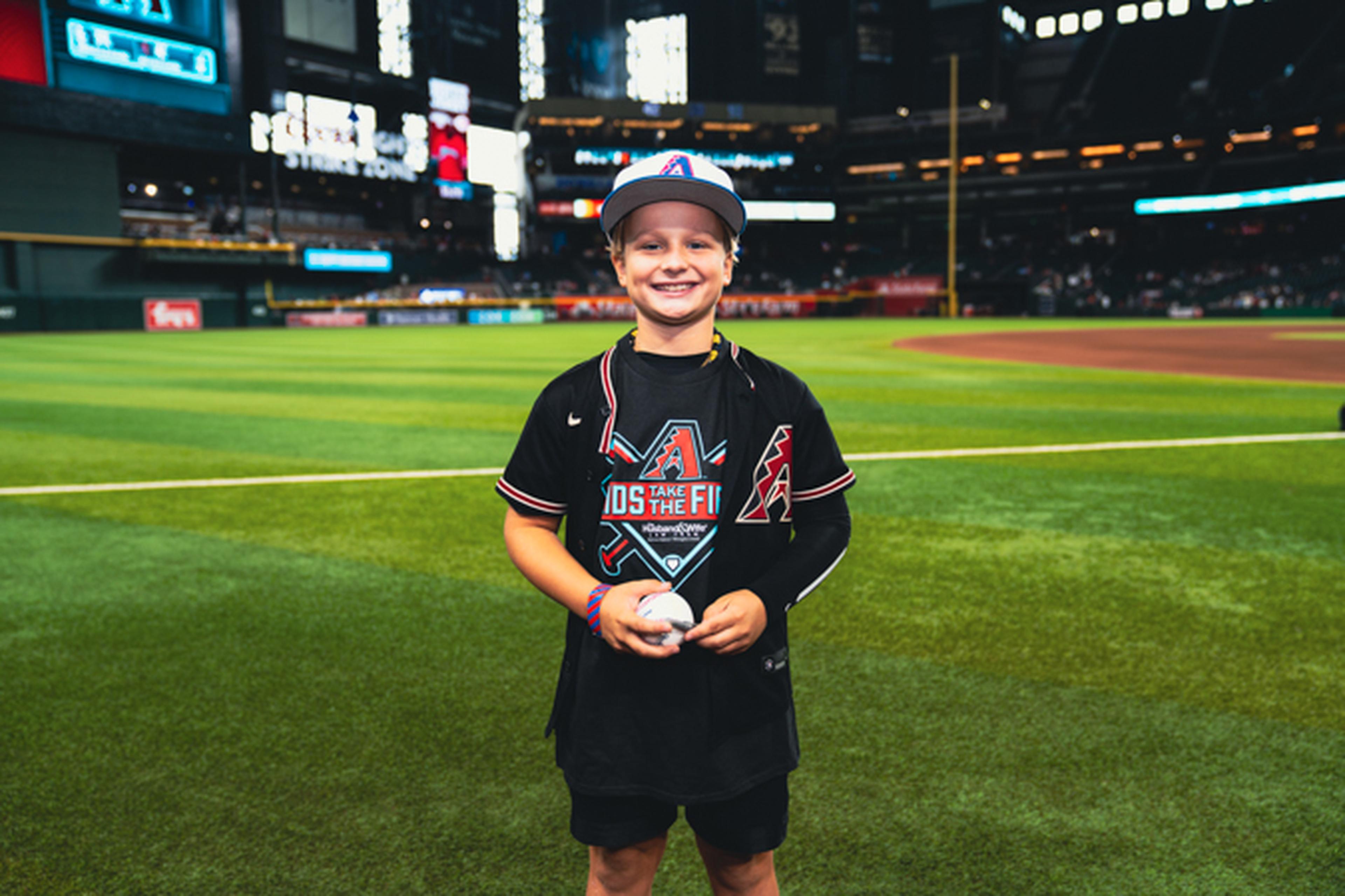Smiling child in a baseball jersey and cap stands on a baseball field holding a ball, with a stadium in the background.