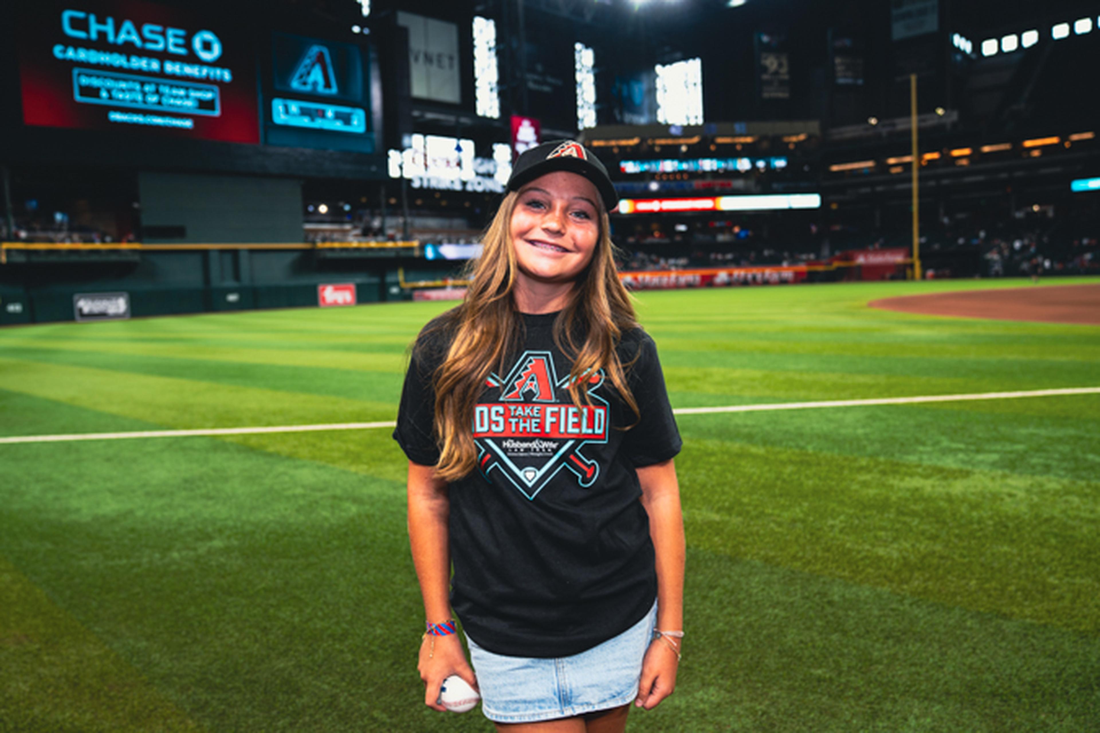 Young person on a baseball field wearing a black cap and shirt with team logo, smiling and holding a baseball. Stadium lights in the background.