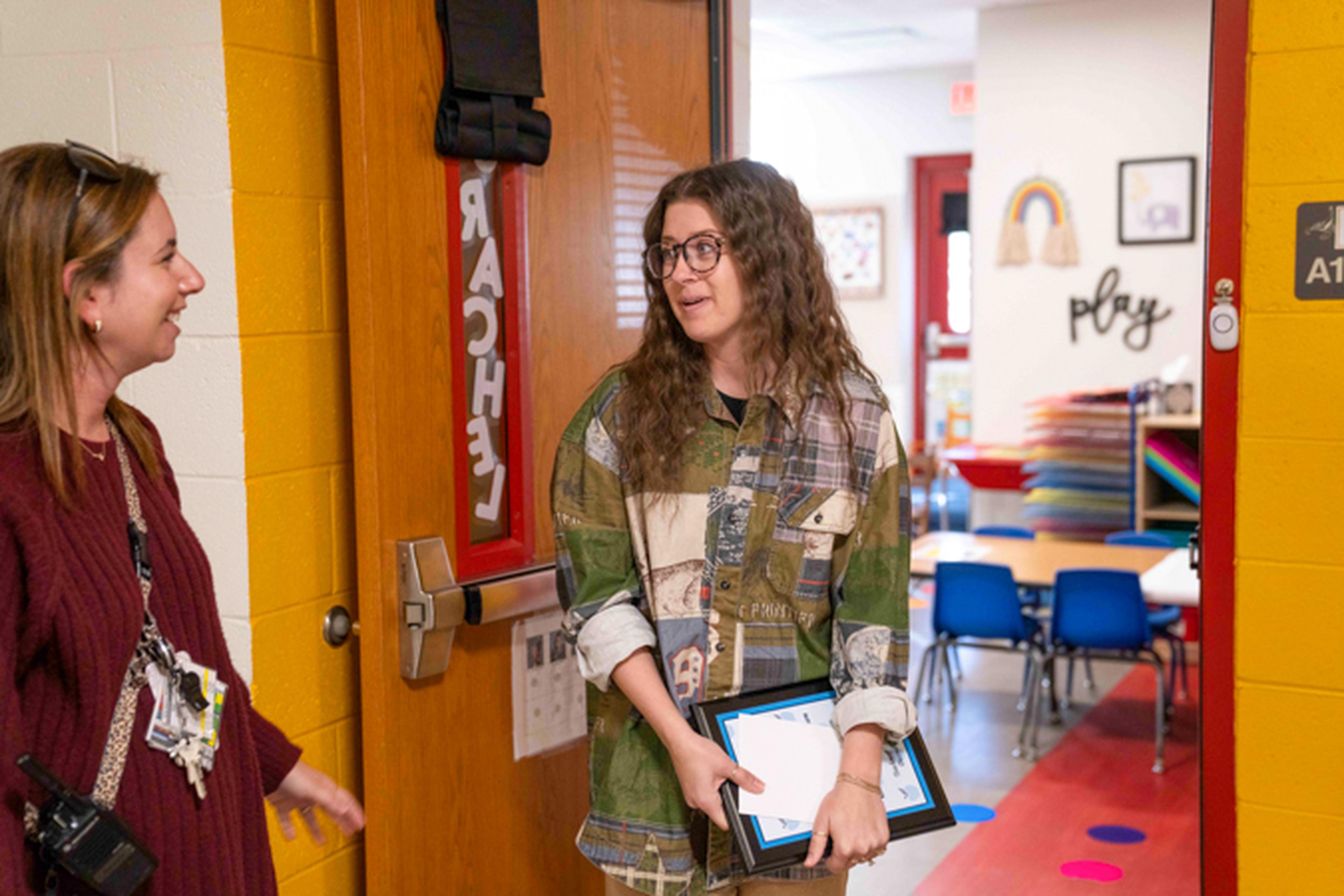 two educators standing in a school doorway, one holding a plaque