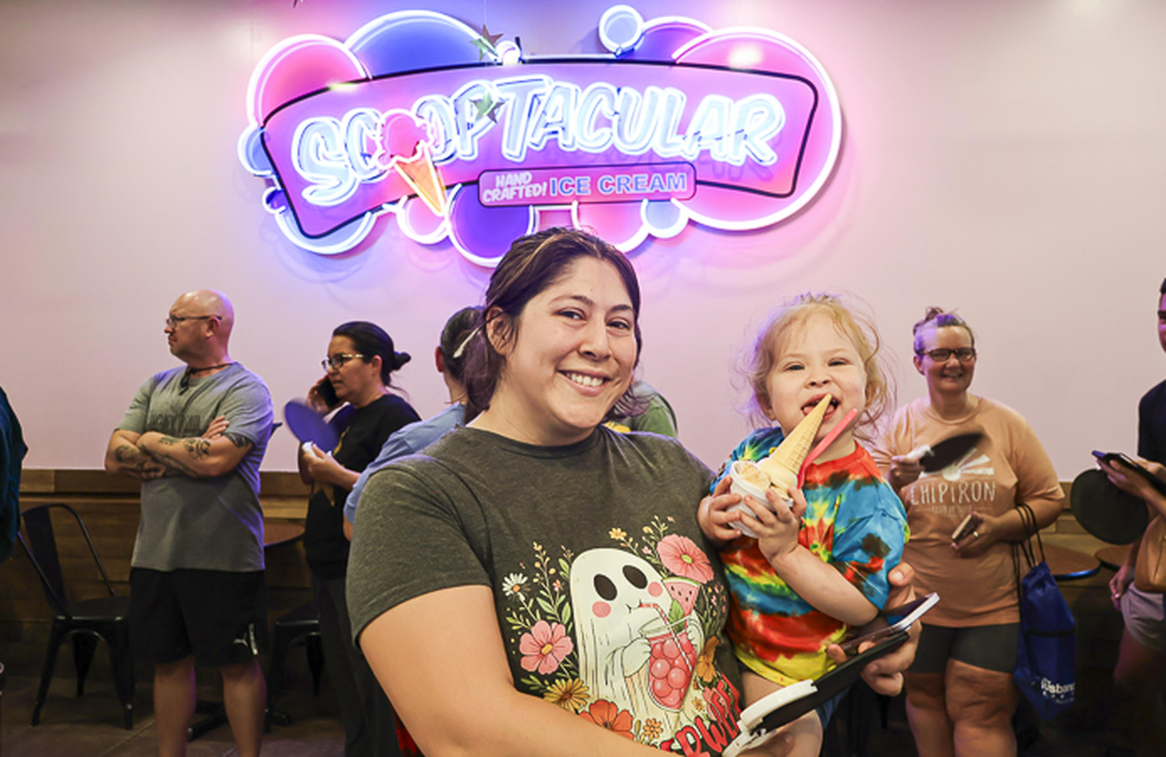A mother and child share a smile in front of the SCOOPTACULAR Handcrafted Ice Cream sign as families gather for a fun-filled community event.