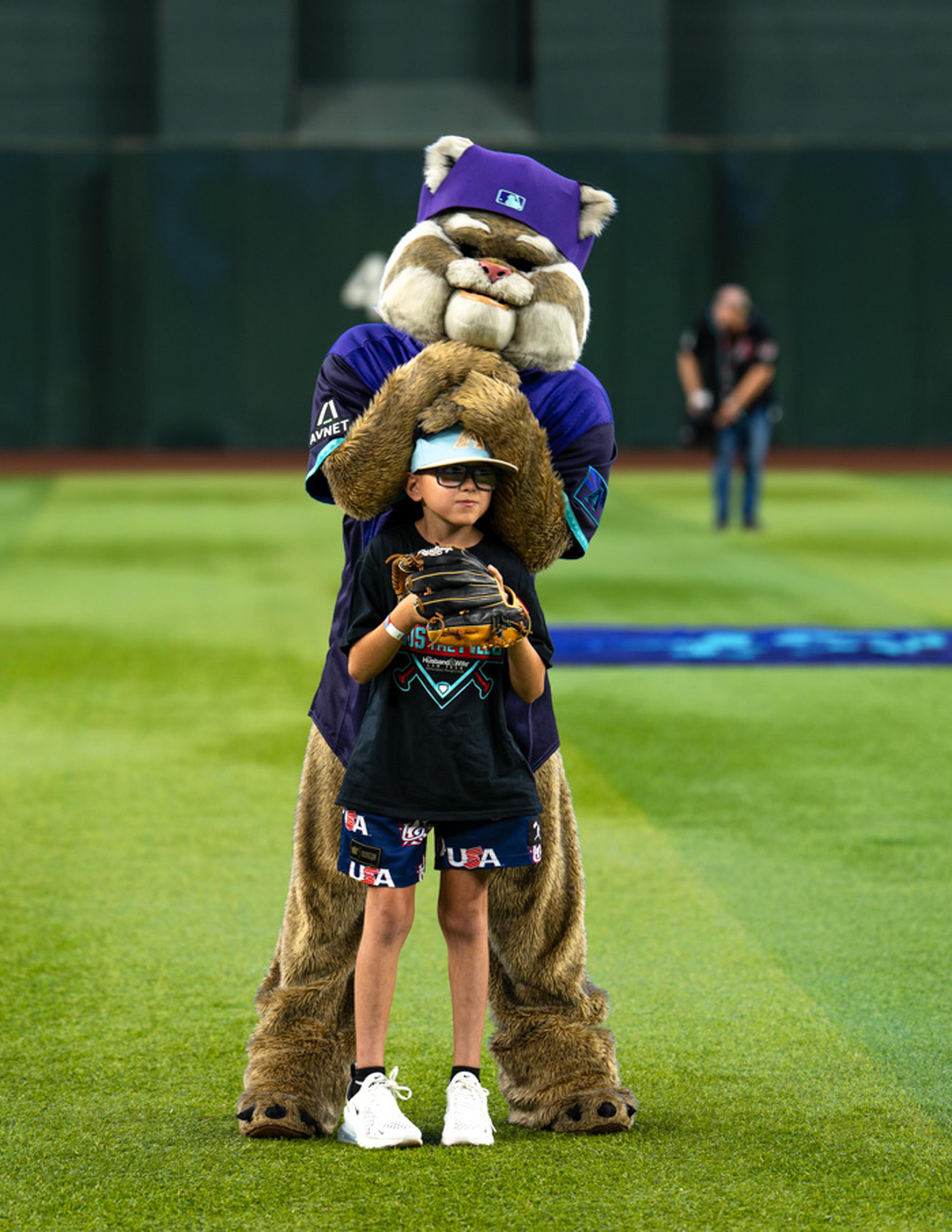 A young Kids Take the Field participant stands ready on the field with his baseball glove while Baxter adds a playful surprise, creating a fun and unforgettable moment at Chase Field.