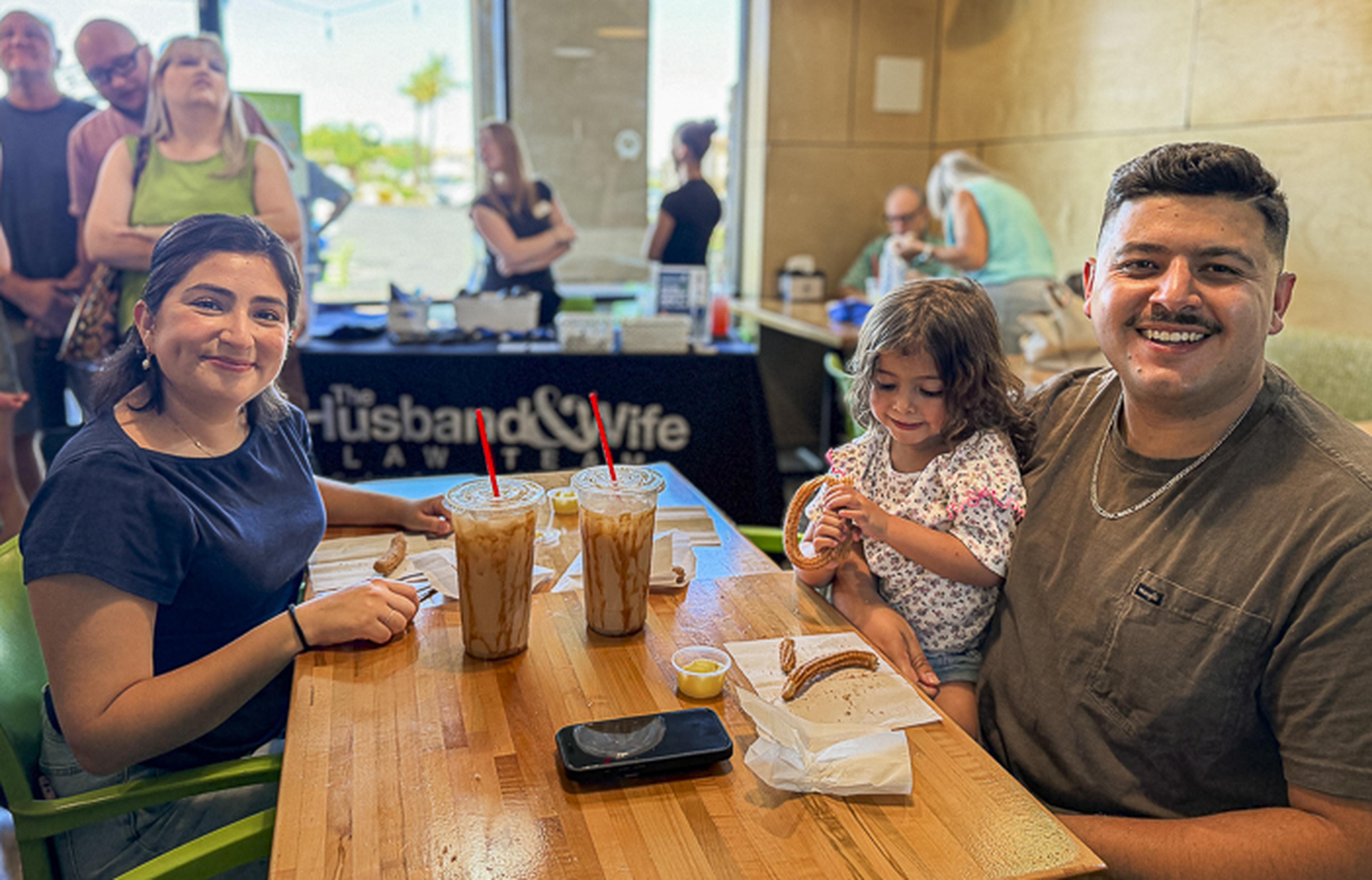 A smiling family sits inside Dulce Churro Café in Gilbert, Arizona, enjoying drinks and pastries with a “Husband & Wife Law Team” banner in the background.