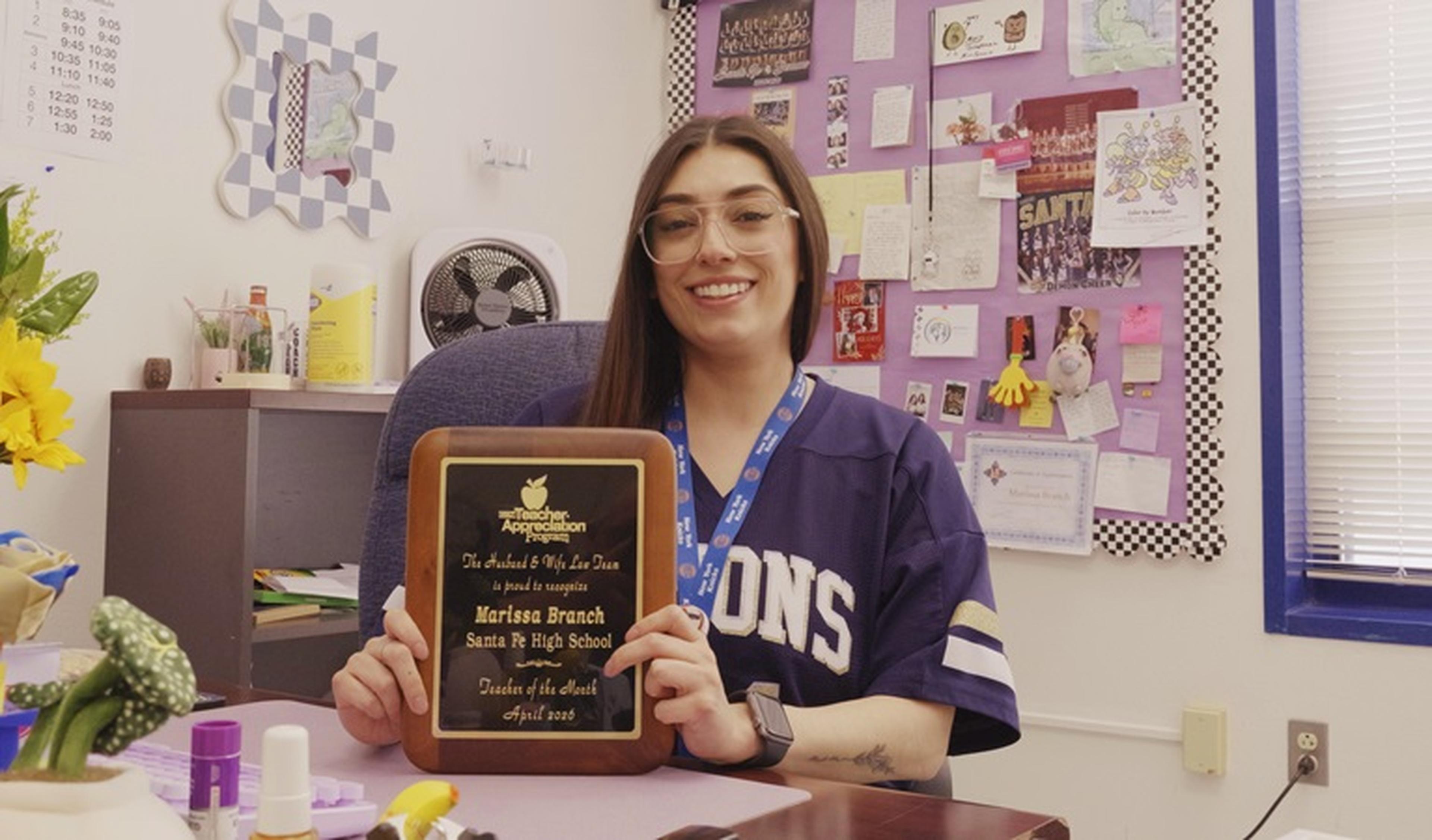 A smiling Ms. Branch holds her “Teacher of the Month” plaque in her classroom, with a bulletin board and desk items behind her.