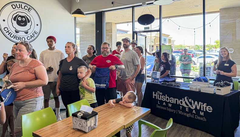 Inside Dulce Churro Café, customers gather near a table featuring a Husband & Wife Law Team banner while a child leans comfortably on the table. A group of customers stands inside Dulce Churro Café beside a table displaying a “Husband & Wife Law Team” banner, with a child resting playfully on the tabletop.