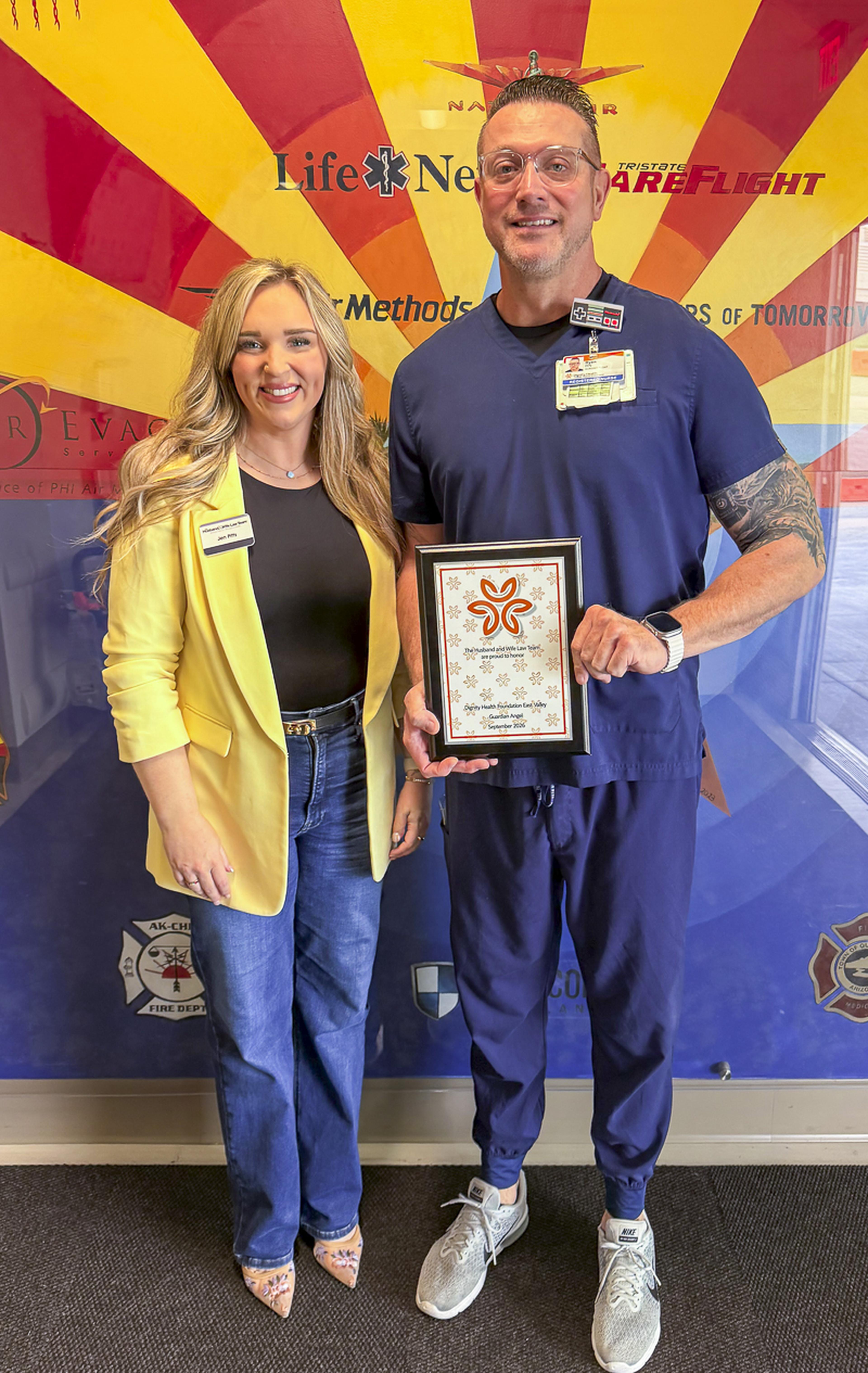 Jen from the Husband & Wife Law Team with nurse Ryan; Ryan holding a framed certificate, standing against a colorful wall with logos and graphics in the background.