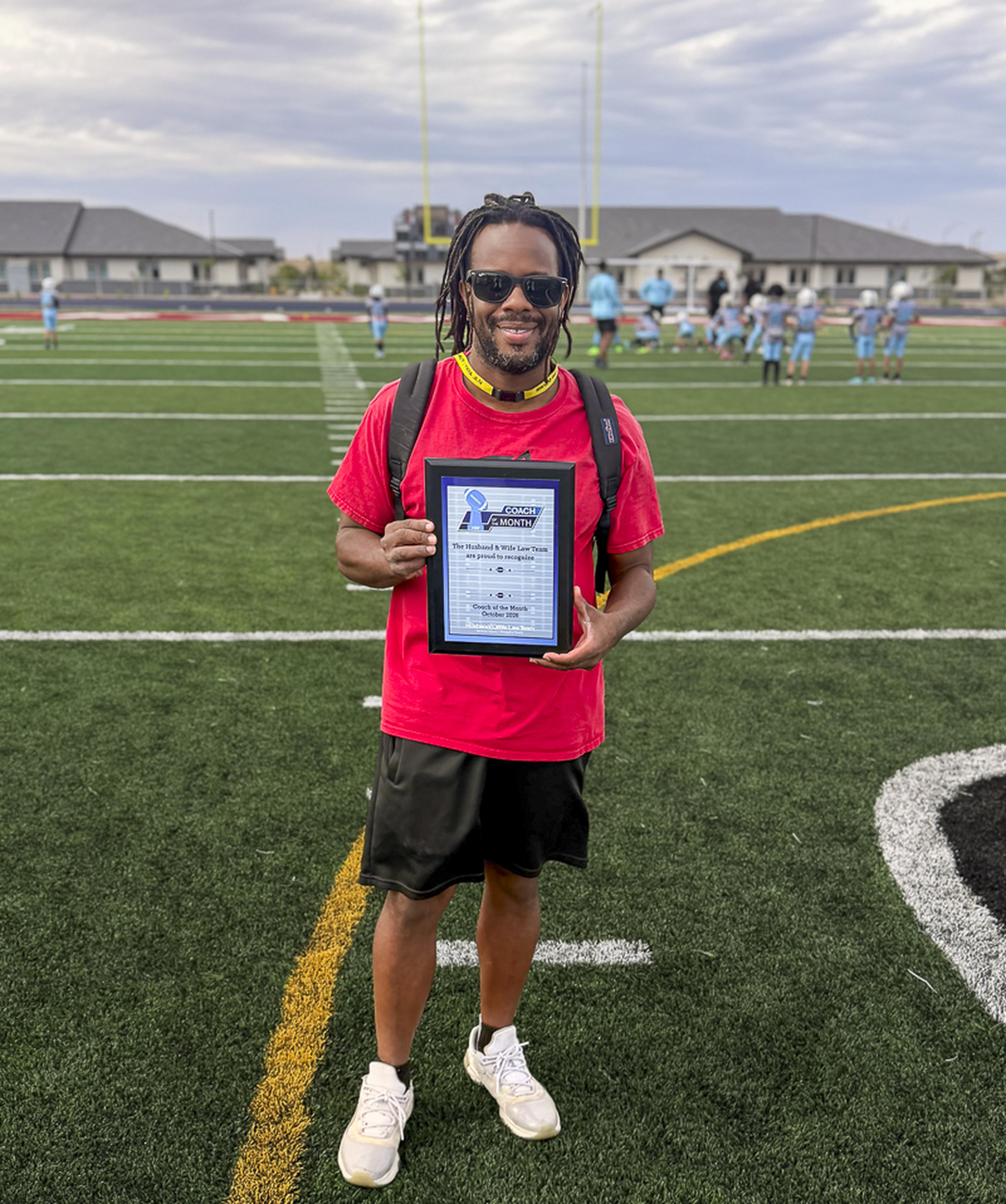 Coach Edwards proudly holds his Husband & Wife Law Team Coach of the Month award on the football field as players gather in the background.