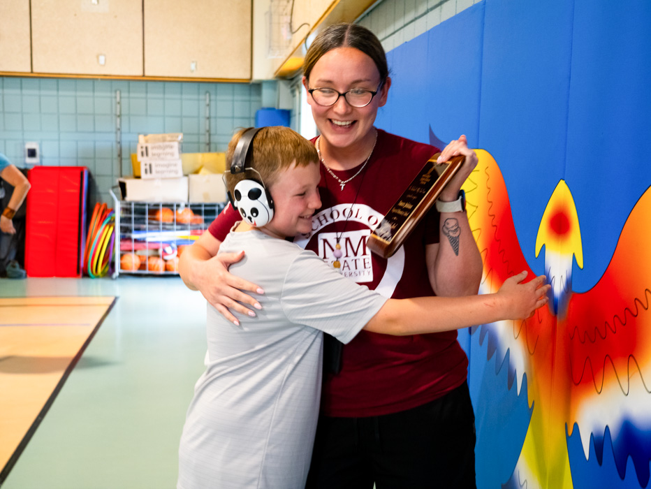 Ms. Quintana holding a Teacher Appreciation Award  holding a plaque hugs a child wearing headphones in a colorful gym setting.