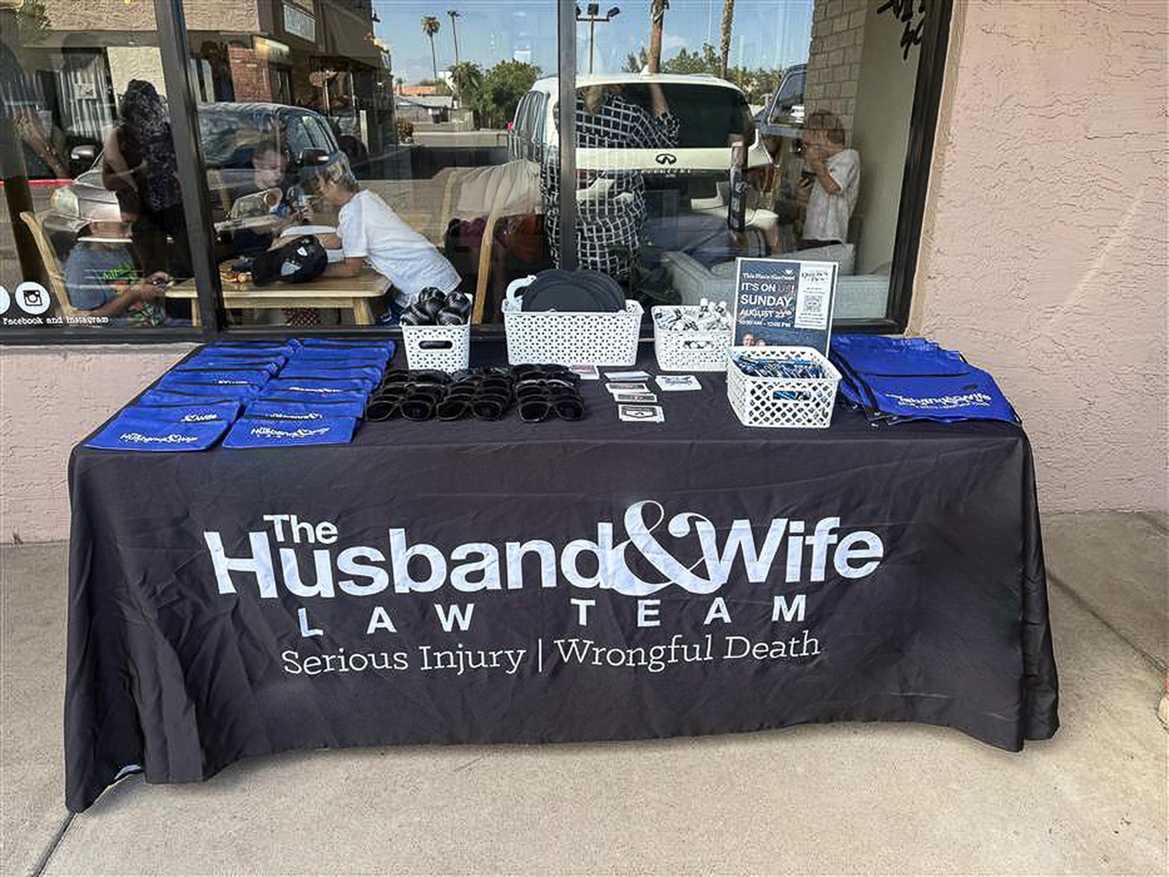 A Husband & Wife Law Team outreach table outside Quiches & Pies Bakery features informational materials, tote bags, and giveaways as part of a community-focused HEART event.