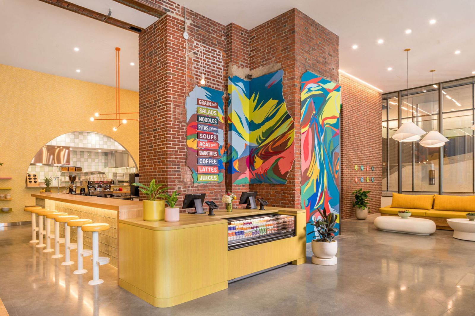 Modern café in Seaport Boston with exposed brick walls, a colorful mural, order counter with cold-pressed juices, beverage bar with stools, menu boards, and cozy seating area with couches.