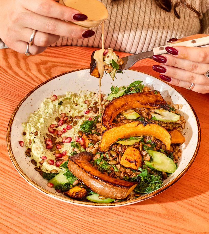 A bowl of roasted koginut squash slices, kale, toasted seeds, and pomegranate atop a spicy jalapeño feta base, with citrus tahini dressing being drizzled by a person.