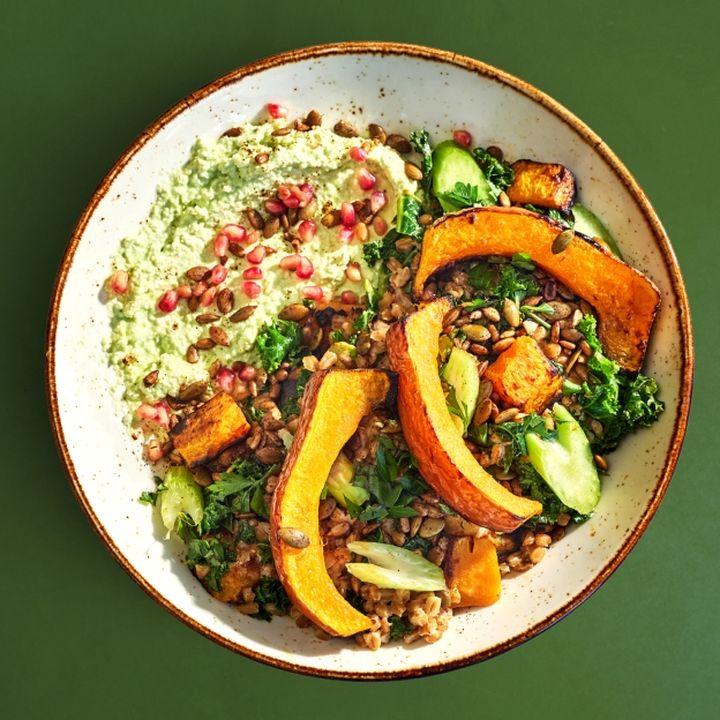 A bowl filled with roasted koginut squash, kale, celery, farro, toasted pumpkin & sunflower seeds, topped with spicy jalapeño feta dip and pomegranate seeds.