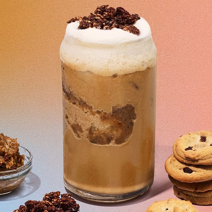 A frothy iced coffee topped with vanilla cold foam and chocolate cookie crumbles beside a stack of chocolate chip cookies and a small dish of granola on a colorful background.