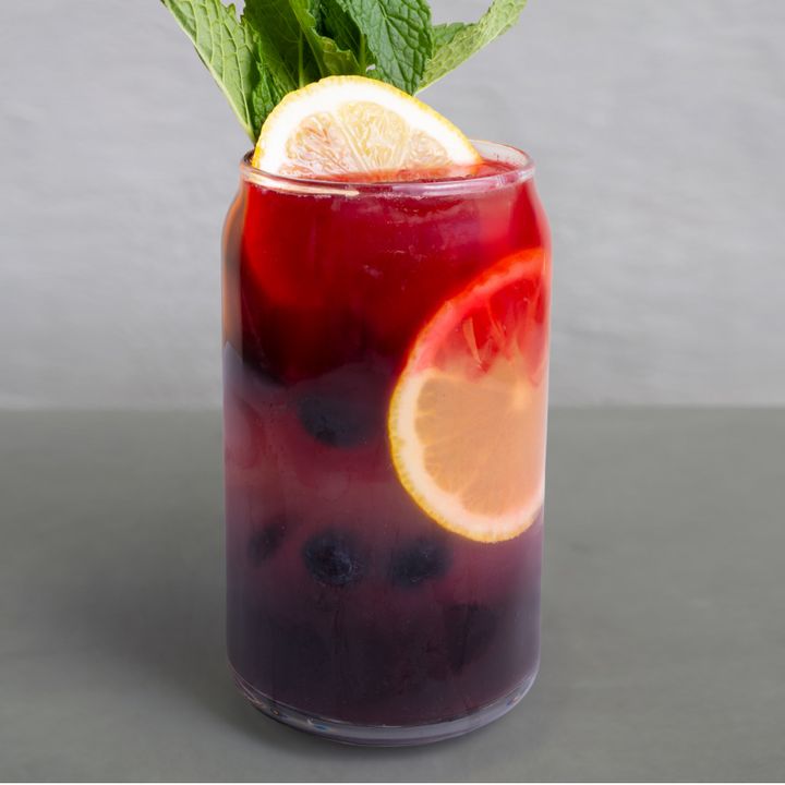A tall glass of dark red iced tea with lemon slices, blueberries, hibiscus and mint leaves on top.