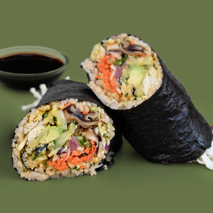 Vegetarian sushi burrito filled with rice, avocado, carrots, mushrooms, and other veggies, served with a small dish of tamari sauce on the side.