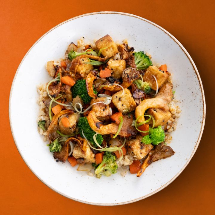 A vibrant dish of steamed vegetables, tofu, and brown rice, garnished with scallions, served on a white plate against an orange background.