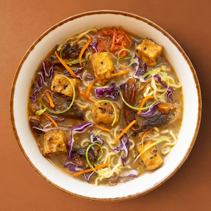 Bowl of ramen with tofu, red cabbage, carrots, mushrooms, and scallions in chickpea miso broth, set on a brown background.