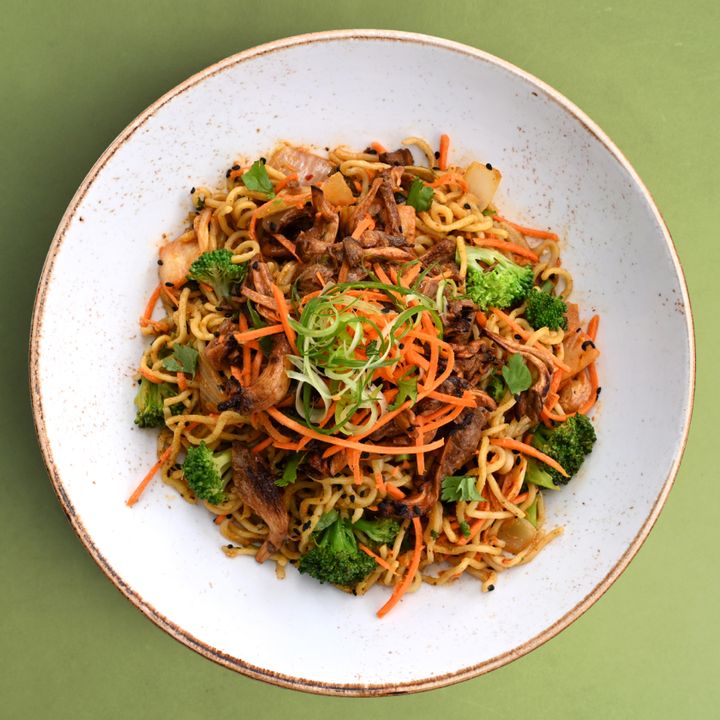 A healthy plate of ramen noodles topped with chili garlic mushrooms, broccoli, shredded carrots, and scallions on a green background.