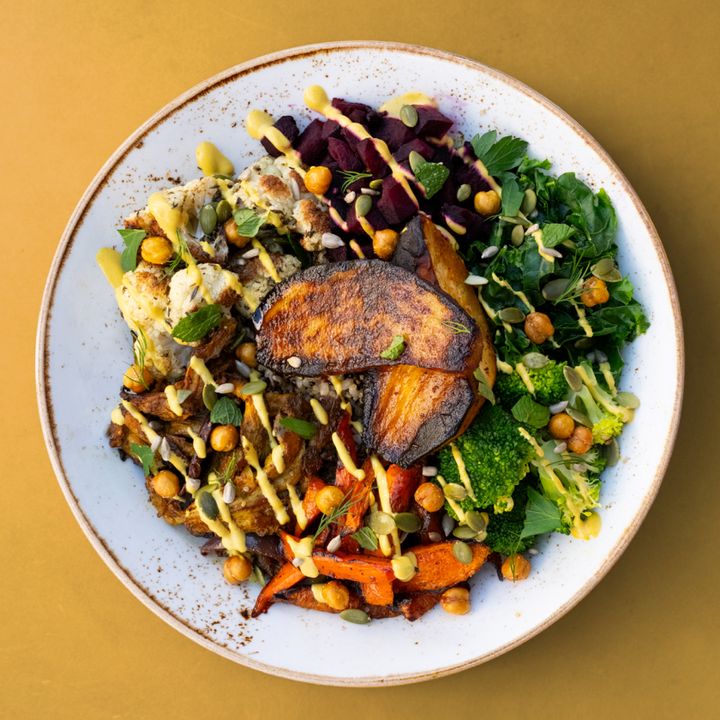 A colorful, healthy rice bowl with roasted white sweet potato and cauliflower, steamed kale, broccoli, carrots and beets, chickpeas and a drizzle of golden tahini sauce on a white plate against a yellow background.