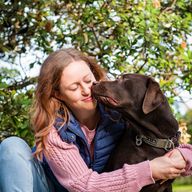 Woman sitting outdoors smiles as she embraces a brown dog. They sit under a tree with green leaves and yellow flowers.