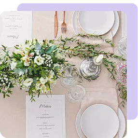 Elegant table setting with white plates, menus, leafy floral centerpiece, glassware, and copper cutlery on a light tablecloth.