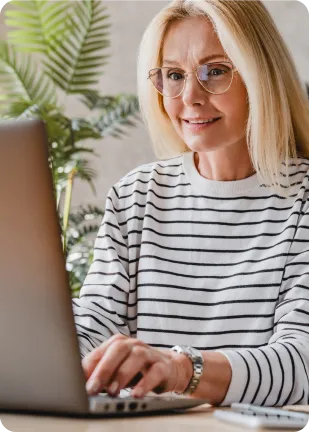 A woman with glasses and a striped sweater using a laptop, sitting at a desk with plants in the background.