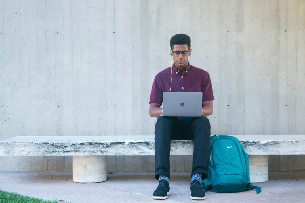 A student in a purple shirt sits with a laptop computer next to a backpack