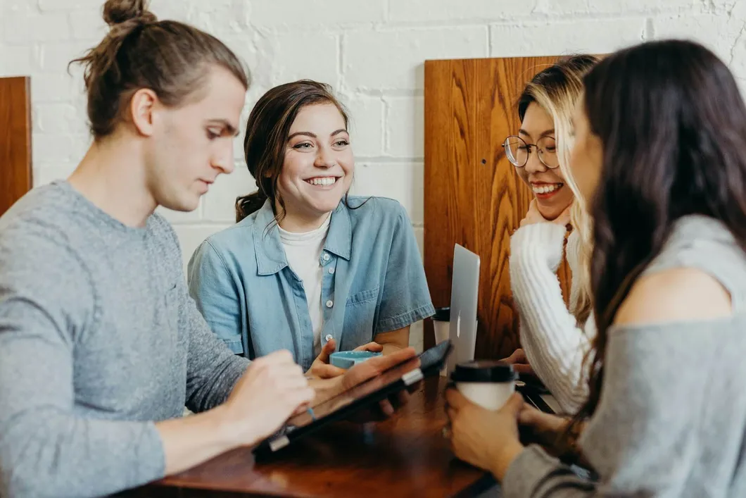 Four young people sitting at a table in a coffee shop, talking, drinking coffee and working on electronics
