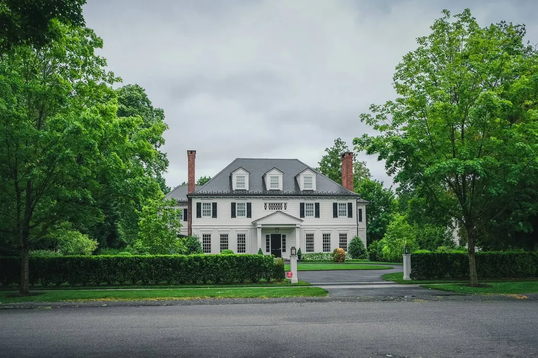 Large two-story white house on a green lawn and green trees under a white sky