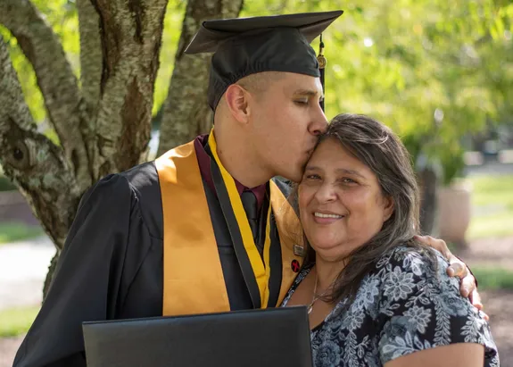 Graduating male student wearing cap and gown, kissing a woman on her forehead