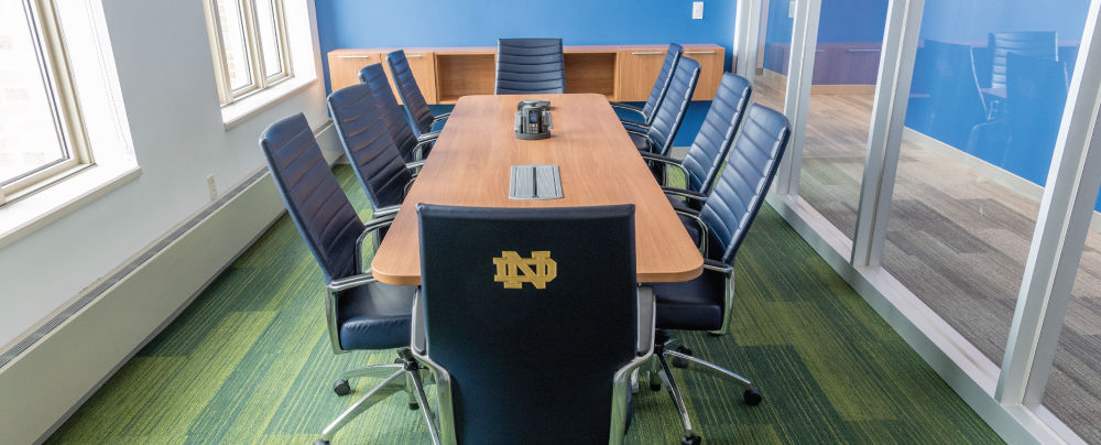 A conference room with a wooden table, blue chairs, and a blue accent wall. The chair at the head of the table has a Notre Dame logo on the back.