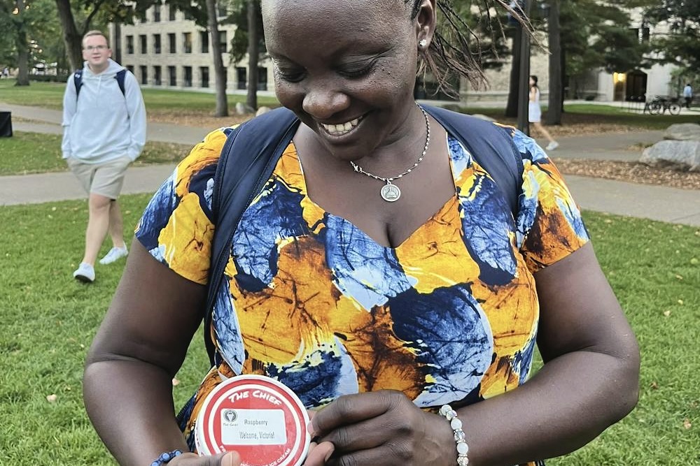 Smiling woman holds a red ice cream pint container labeled "Raspberry, Welcome Victoria!" on a grassy area with trees and buildings in the background.