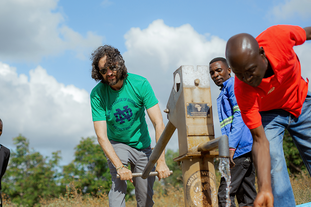 A man in a green shirt pumps water from a well in Malawi, assisted by others, under a partly cloudy sky.