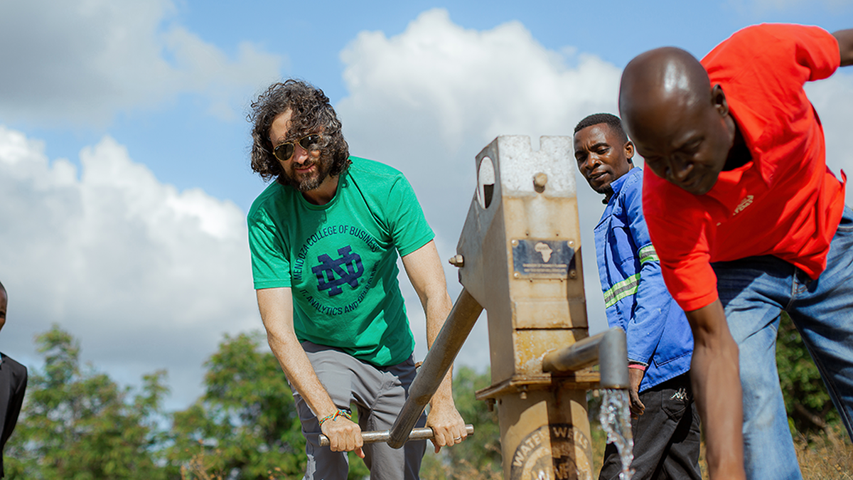 A man in a green shirt pumps water from a well in Malawi, assisted by others, under a partly cloudy sky.