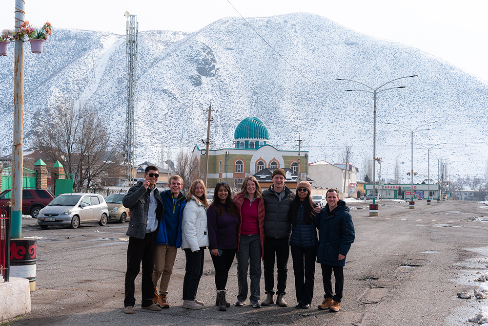 KZ BOTFL 2025 A group of Notre Dame students and Kyrgyz partners stands together on a snowy street with mountains and a mosque with a teal dome in the background.