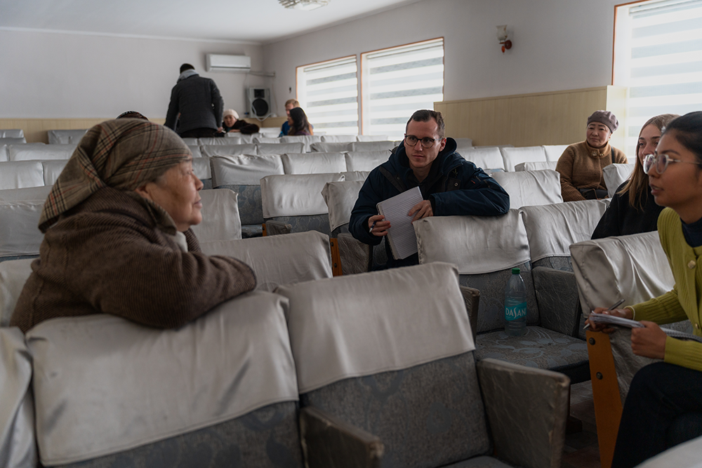 Notre Dame MBA students engage in conversation with a Kyrgyz entrepreneur and an interpreter in a room with rows of empty seats. Some hold papers, and one person wears a headscarf.