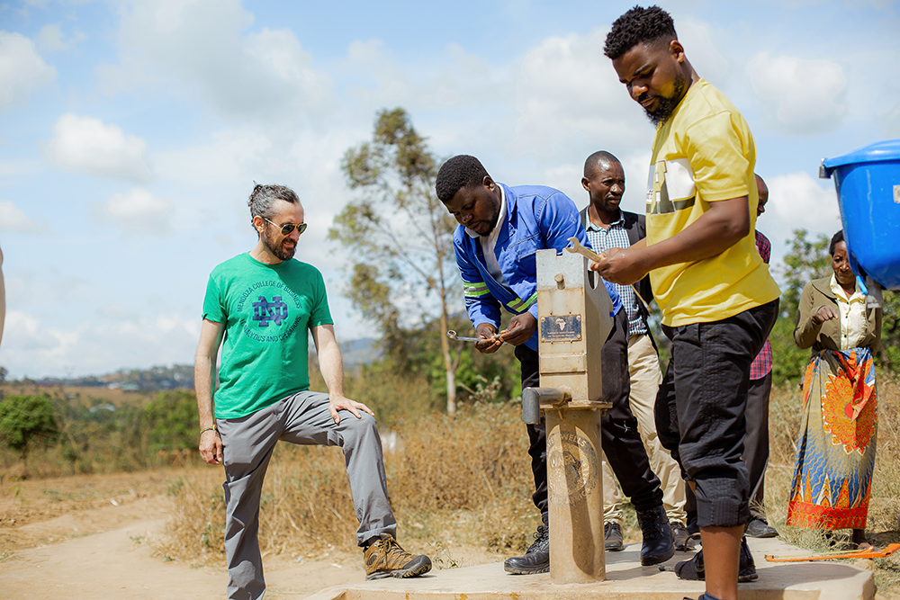 A group of people gathers around a manual water pump in rural Malawi with one person repairing the pump. Trees and a cloudy sky are in the background.