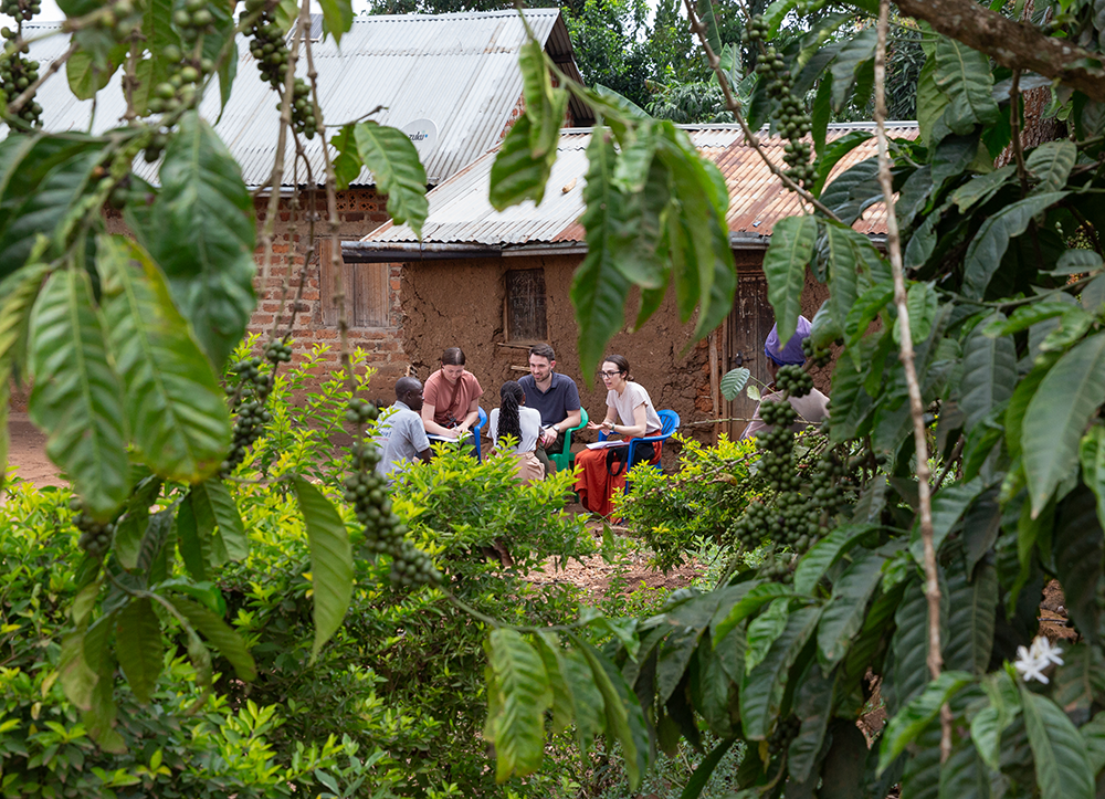 Five people sit in a circle outside brick mud huts with metal roofs amidst greenery in Uganda.