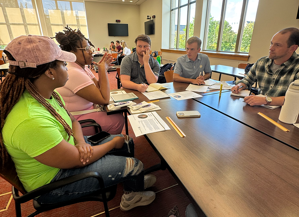 A group of five people sit around a table in an MBA classroom at Notre Dame, engaged in discussion with papers, notebooks, and phones on the table.