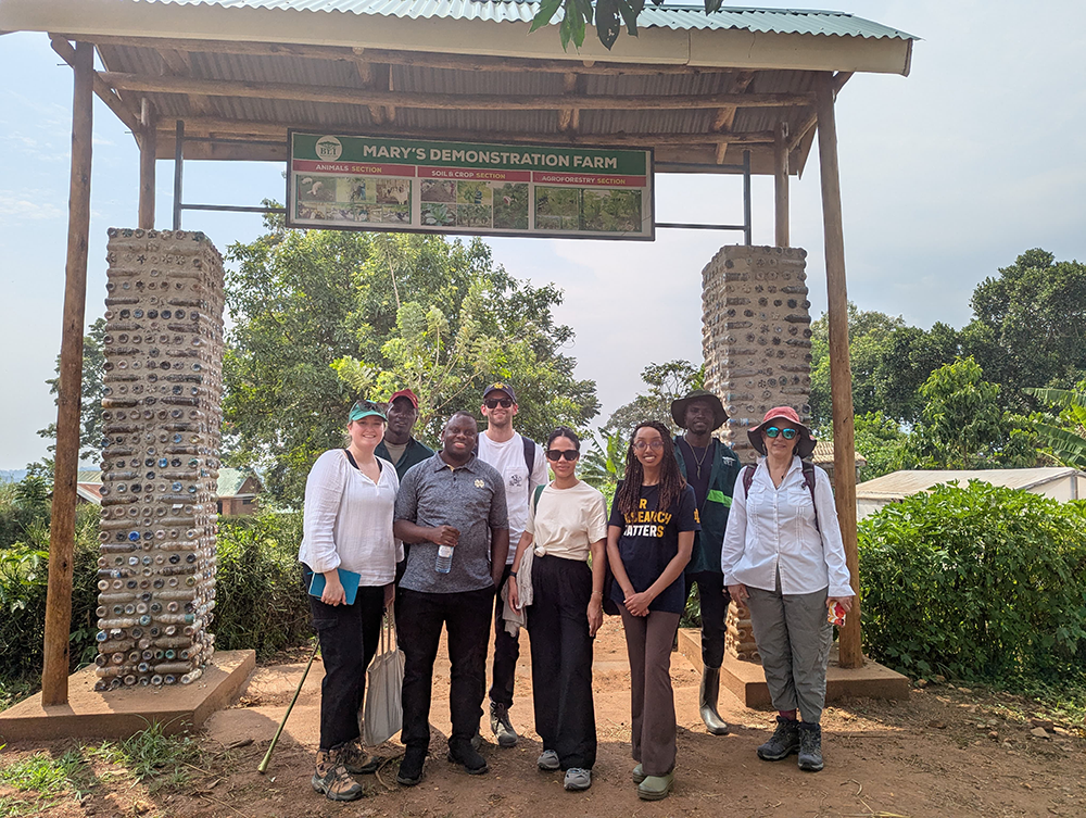 In rural Uganda, group of people stands under a sign reading "Mary's Demonstration Farm," surrounded by greenery and a structure made of bottles.