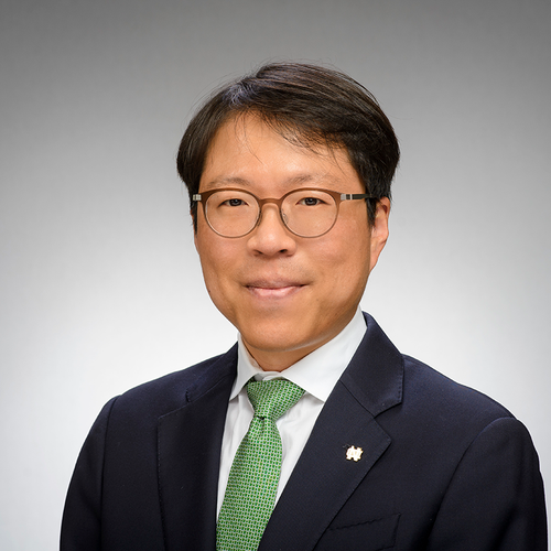 A man in a suit with glasses and a green tie, smiling against a plain background.